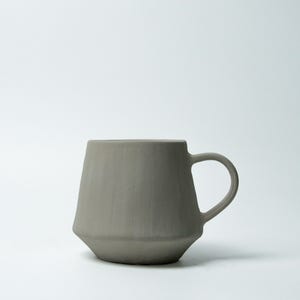 K&ouml;nnte beinhalten: Eine graue Keramik-Tasse mit einem einzigartigen, geometrischen Design. Die Tasse hat einen zylindrischen Henkel und einen leicht konischen K&ouml;rper, was ihr eine moderne &Auml;sthetik verleiht. Die neutrale Farbe und die schlichte Form machen sie f&uuml;r verschiedene Getr&auml;nke geeignet.