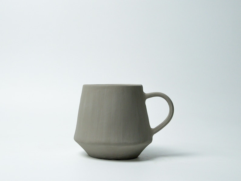 K&ouml;nnte beinhalten: Eine graue Keramik-Tasse mit einem einzigartigen, geometrischen Design. Die Tasse hat einen zylindrischen Henkel und einen leicht konischen K&ouml;rper, was ihr eine moderne &Auml;sthetik verleiht. Die neutrale Farbe und die schlichte Form machen sie f&uuml;r verschiedene Getr&auml;nke geeignet.