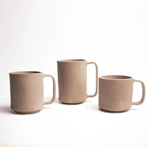 May include: Three beige ceramic mugs of varying sizes are arranged on a white background. Each mug has a simple, cylindrical design with a handle. The mugs appear to be handmade and have a matte finish.
