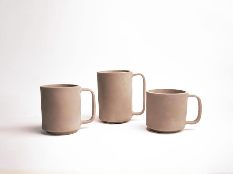May include: Three beige ceramic mugs of varying sizes are arranged on a white background. Each mug has a simple, cylindrical design with a handle. The mugs appear to be handmade and have a matte finish.