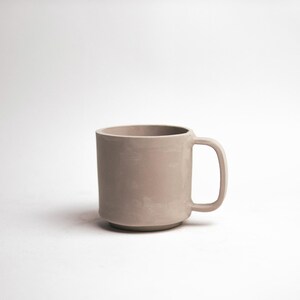 May include: A neutral-toned ceramic mug with a rounded base and a simple handle. The mug has a smooth, matte finish and a cylindrical shape. The mug is designed for hot or cold beverages.