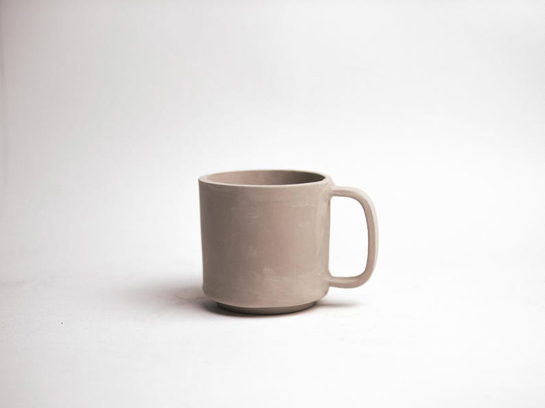 May include: A neutral-toned ceramic mug with a rounded base and a simple handle. The mug has a smooth, matte finish and a cylindrical shape. The mug is designed for hot or cold beverages.