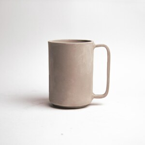 May include: A tall, cylindrical mug in a muted taupe color. The mug has a simple, modern design with a rectangular handle. The mug is approximately 6 inches tall and is suitable for coffee, tea, or other beverages.