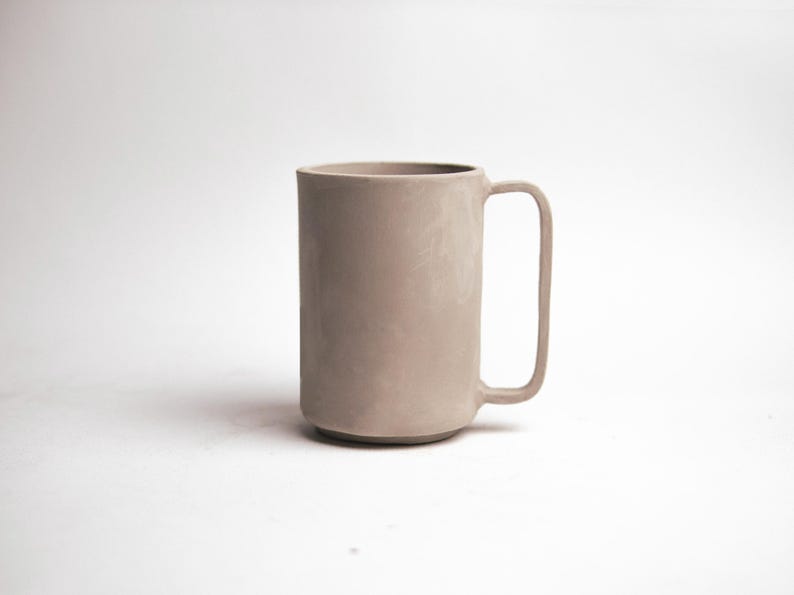 May include: A tall, cylindrical mug in a muted taupe color. The mug has a simple, modern design with a rectangular handle. The mug is approximately 6 inches tall and is suitable for coffee, tea, or other beverages.