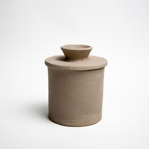 May include: A cylindrical, light brown ceramic butter dish with a matching lid. The lid has a small, rounded handle on top. The dish is designed to hold butter and keep it fresh. The butter dish is a neutral color and has a simple, minimalist design.