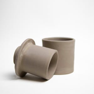 May include: Two gray ceramic pieces, one cylindrical and the other with a flared base, sit against a white background. The cylindrical piece stands upright, while the other rests on its side. The objects appear to be handmade pottery.