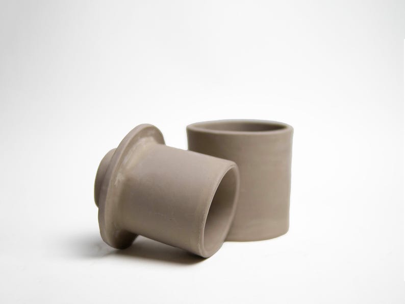 May include: Two gray ceramic pieces, one cylindrical and the other with a flared base, sit against a white background. The cylindrical piece stands upright, while the other rests on its side. The objects appear to be handmade pottery.