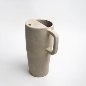 K&ouml;nnte beinhalten: Eine hohe, beige Keramiktasse mit einem einzigartigen Design. Die Tasse hat einen Henkel und eine teilweise abgedeckte Oberseite mit einem kleinen Loch. Die Form der Tasse ist zylindrisch, mit einem leicht verj&uuml;ngten Boden.