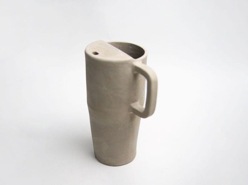 K&ouml;nnte beinhalten: Eine hohe, beige Keramiktasse mit einem einzigartigen Design. Die Tasse hat einen Henkel und eine teilweise abgedeckte Oberseite mit einem kleinen Loch. Die Form der Tasse ist zylindrisch, mit einem leicht verj&uuml;ngten Boden.