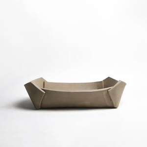 May include: A beige, rectangular tray with a folded, geometric design. The tray has a shallow depth and is made of a solid, matte material. The corners are angled, and the sides slope inward. The tray is suitable for holding small items.