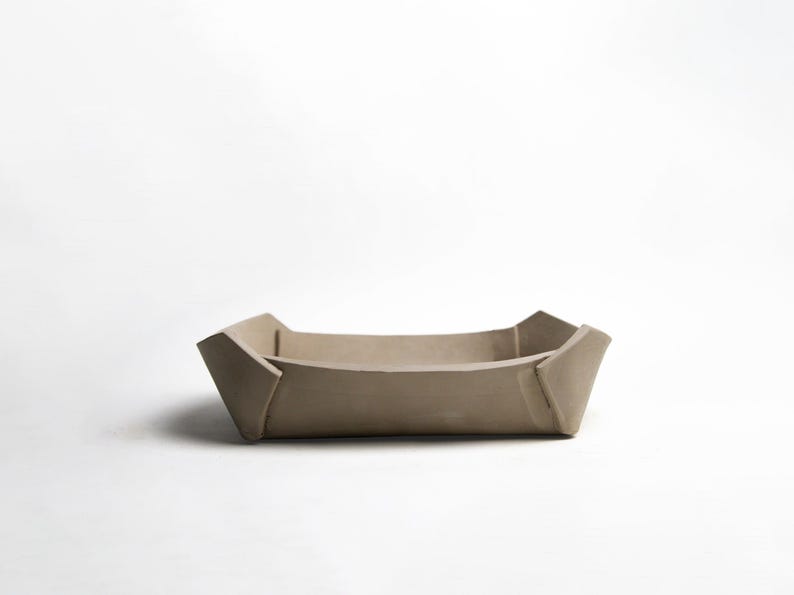 May include: A beige, rectangular tray with a folded, geometric design. The tray has a shallow depth and is made of a solid, matte material. The corners are angled, and the sides slope inward. The tray is suitable for holding small items.