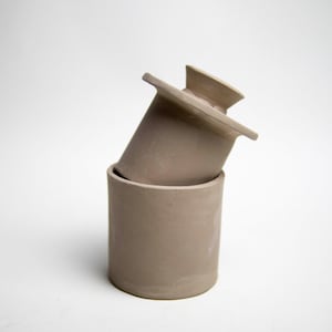 May include: A two-piece, light brown ceramic butter dish. The base is a cylindrical container, and the lid is rectangular with a handle. The lid is resting on the base at an angle, with a white background.