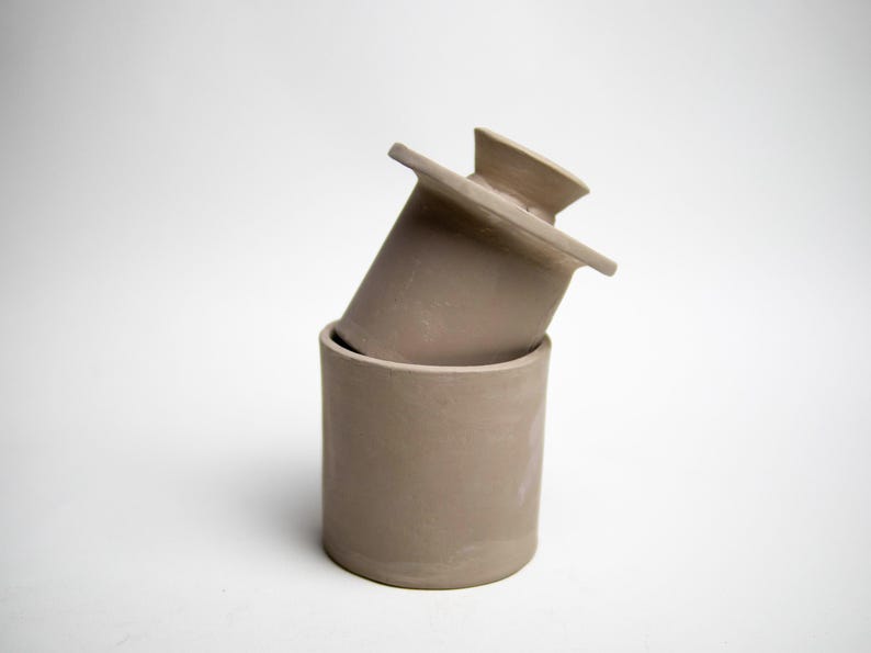 May include: A two-piece, light brown ceramic butter dish. The base is a cylindrical container, and the lid is rectangular with a handle. The lid is resting on the base at an angle, with a white background.