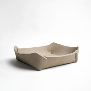 May include: A beige ceramic tray, boat-shaped, with a rectangular base and angled sides. The corners are pointed. The neutral colour and minimalist design suggest a modern aesthetic, suitable for home decor or serving.