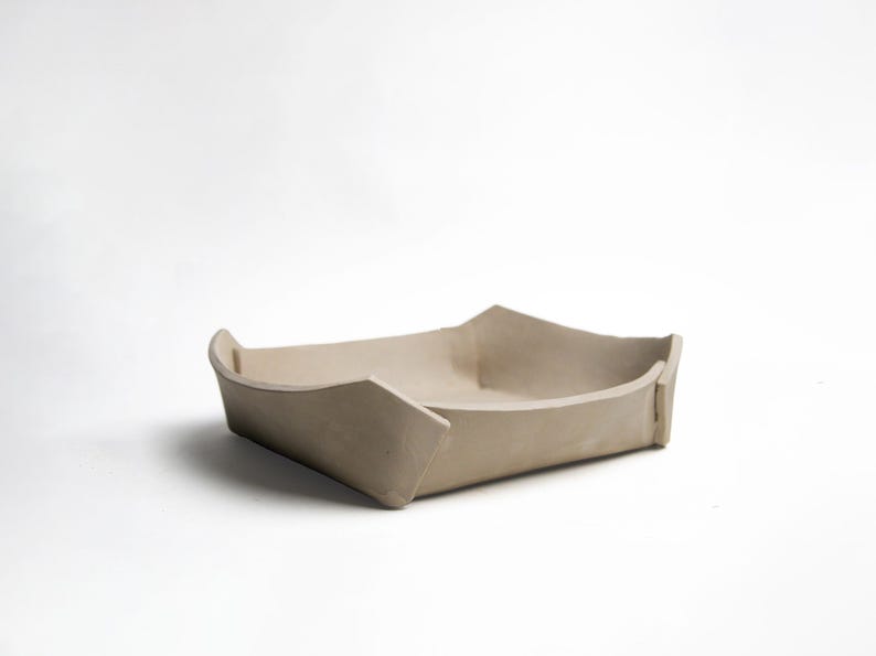 May include: A beige ceramic tray, boat-shaped, with a rectangular base and angled sides. The corners are pointed. The neutral colour and minimalist design suggest a modern aesthetic, suitable for home decor or serving.