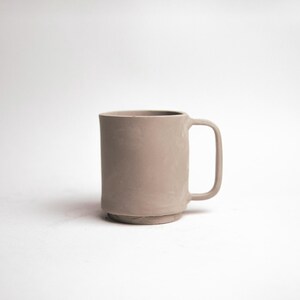 May include: A neutral-toned ceramic mug with a cylindrical body and a square handle. The mug has a slightly textured surface and sits on a small, raised base. The mug is a light, muted brown color.