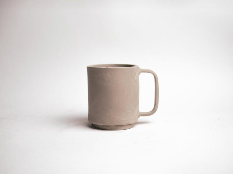 May include: A neutral-toned ceramic mug with a cylindrical body and a square handle. The mug has a slightly textured surface and sits on a small, raised base. The mug is a light, muted brown color.