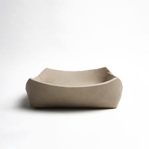 May include: A square, beige ceramic bowl with softly curved sides. The bowl has a smooth, matt finish and is set against a plain white background. The design is minimalist and modern, suitable for serving or display.