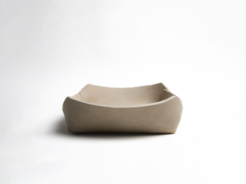 May include: A square, beige ceramic bowl with softly curved sides. The bowl has a smooth, matt finish and is set against a plain white background. The design is minimalist and modern, suitable for serving or display.