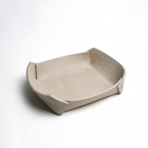 May include: A beige ceramic tray with a rectangular shape and slightly raised sides. The tray has a minimalist design with clean lines and a matte finish. The corners are folded and have a small vertical cut. Suitable for serving or display.