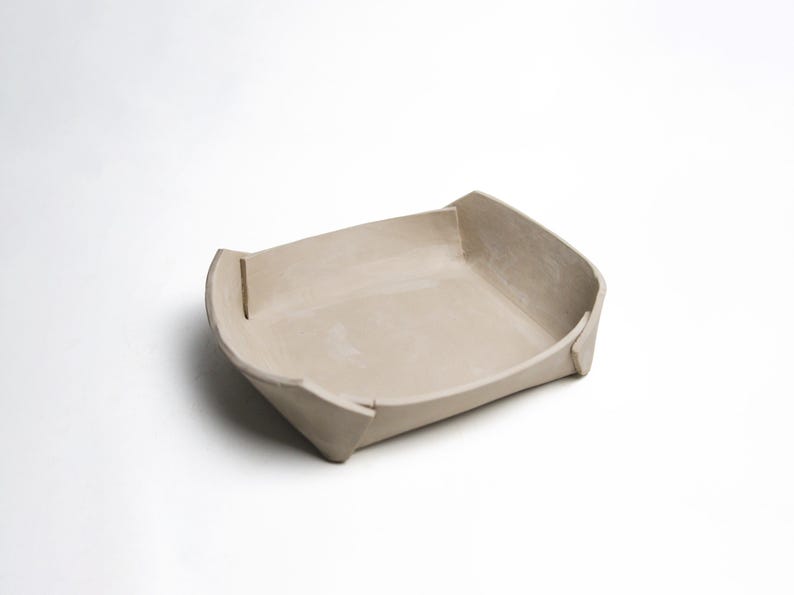 May include: A beige ceramic tray with a rectangular shape and slightly raised sides. The tray has a minimalist design with clean lines and a matte finish. The corners are folded and have a small vertical cut. Suitable for serving or display.