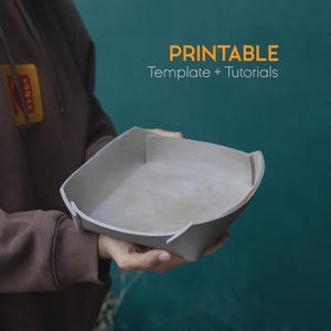 Op de afbeelding: Een vierkante, lichtgrijze bak met gebogen randen wordt vastgehouden. De bak heeft een lichtgekleurd interieur. Oranje tekst luidt "PRINTABLE Template + Tutorials" tegen een teal achtergrond.