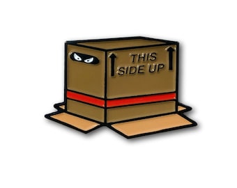 May include: A brown box-shaped enamel pin with a red stripe and the words "THIS SIDE UP." The box has a ninja mask design and black arrows pointing up. The box flaps are open.
