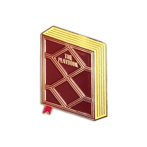Op de afbeelding: Een gouden en rode emaille pin in de vorm van een gesloten boek met de tekst "THE PLAYBOOK" op de voorkant.