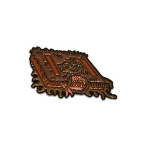 May include: A brown and orange enamel pin depicting a book with a monster's mouth emerging from the pages. The monster has sharp teeth and a pink tongue.