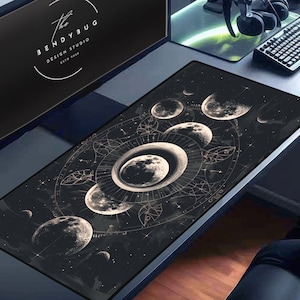 May include: A black and gold mouse pad with a celestial design featuring moons and stars. The design is reminiscent of a constellation chart. The mouse pad is on a desk with a computer monitor, keyboard, and headphones.