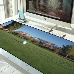 May include: A rectangular desk mat featuring a golf course scene with a golf ball and flag. The mat has a green field, blue sky, and a mountain range in the background. The mat is on a white desk.