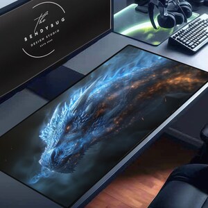 May include: A blue and orange dragon with glowing eyes is featured on a black gaming mouse pad. The mouse pad is on a desk with a computer monitor, headphones, and a keyboard.