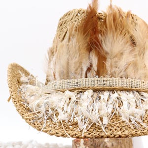 May include: A woven straw hat with a wide brim, adorned with tan feathers and a decorative band. The hat features a cream-colored fringe around the brim, adding a touch of elegance. This hat is perfect for a bohemian or festival look.