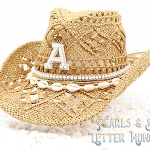 May include: A straw cowboy hat with a pearl and shell trim and a rhinestone letter "A" on the crown. The hat is on a white background with white shells.