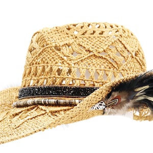 May include: A wide-brimmed straw hat with a crocheted design and a black band with rhinestones. The hat has a feather accent and is a light brown color.