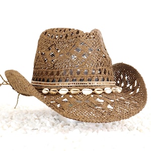 May include: A brown woven cowboy hat with a decorative band of small seashells and a woven design. The hat has a wide brim and a classic cowboy hat shape, perfect for sun protection.