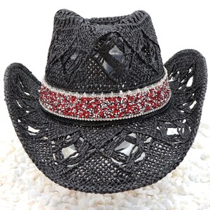 May include: A black cowboy hat with a decorative band of red and silver rhinestones. The hat has a woven design and a wide brim. The hat is sitting on a bed of white stones.
