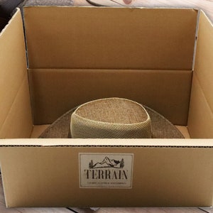 May include: A brown cardboard box with the word "TERRAIN" printed on the side. The box is open and a brown hat is inside.