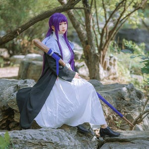 Könnte beinhalten: Eine Person in einem Cosplay-Kostüm mit einem langen weißen Kleid, einem schwarzen und grauen Mantel und einer lila Perücke. Die Person hält einen Holzstab mit blauen Akzenten und sitzt auf einem Felsen. Der Hintergrund zeigt Bäume und Felsen.