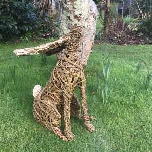 May include: A woven willow sculpture of a rabbit sitting on a bed of green grass. The rabbit is made of natural brown willow branches and has a long, floppy ear.