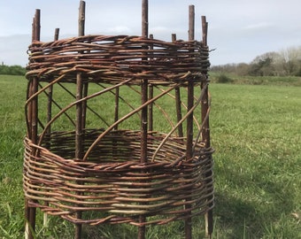 May include: A woven willow basket with a circular base and a cylindrical shape. The basket is made of brown willow branches and is standing on a wooden surface.