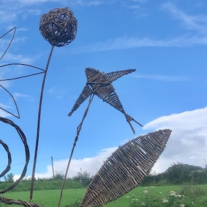 May include: A whimsical garden sculpture made of woven branches, featuring a large round sphere, a star-shaped form, and a long leaf-like shape. The sculpture is set against a blue sky with white clouds.