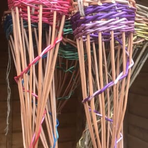 May include: Five hanging baskets made of woven willow branches. The baskets are dyed in various colours, including pink, purple, green, and natural. The baskets are tied together at the top with twine.