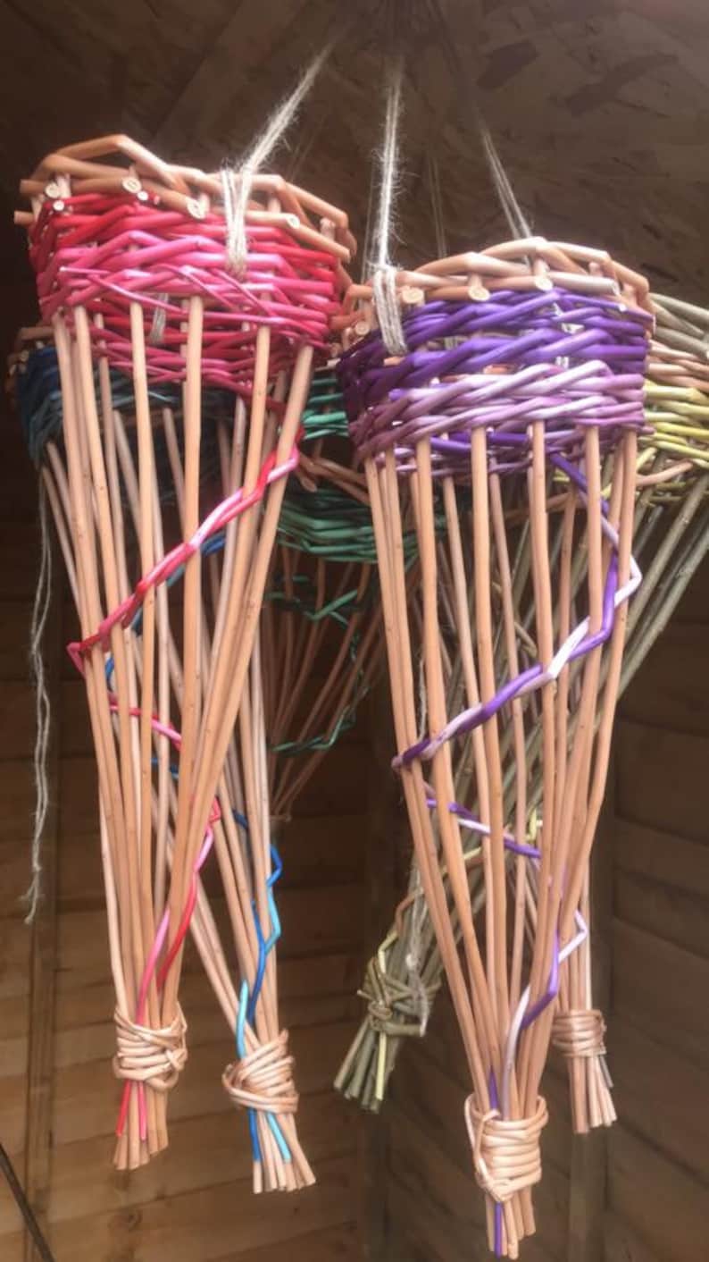 May include: Five hanging baskets made of woven willow branches. The baskets are dyed in various colours, including pink, purple, green, and natural. The baskets are tied together at the top with twine.