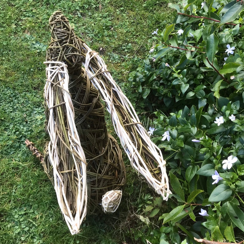 May include: A woven willow rabbit sculpture with white accents. The sculpture is standing on a bed of green grass with white flowers in the background.