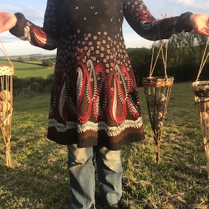 May include: Three woven baskets hanging from twine, each filled with round, light brown objects. The baskets are cone-shaped and have a woven pattern. A person is holding the baskets in front of a green field.