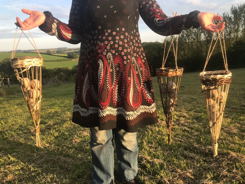 May include: Three woven baskets hanging from twine, each filled with round, light brown objects. The baskets are cone-shaped and have a woven pattern. A person is holding the baskets in front of a green field.