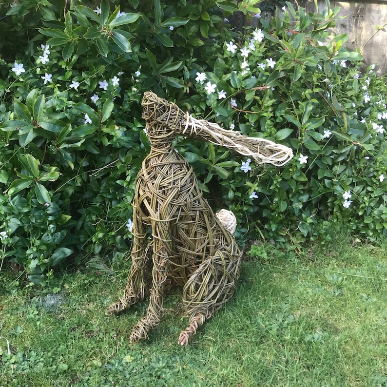 May include: A woven willow sculpture of a hare, with a long, floppy ear, sitting on a bed of green grass. The hare is made of natural materials and has a rustic, handmade look.