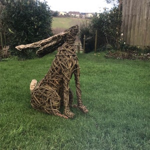 May include: A woven willow sculpture of a rabbit, standing on a grassy lawn. The rabbit is looking up and has its ears perked up.