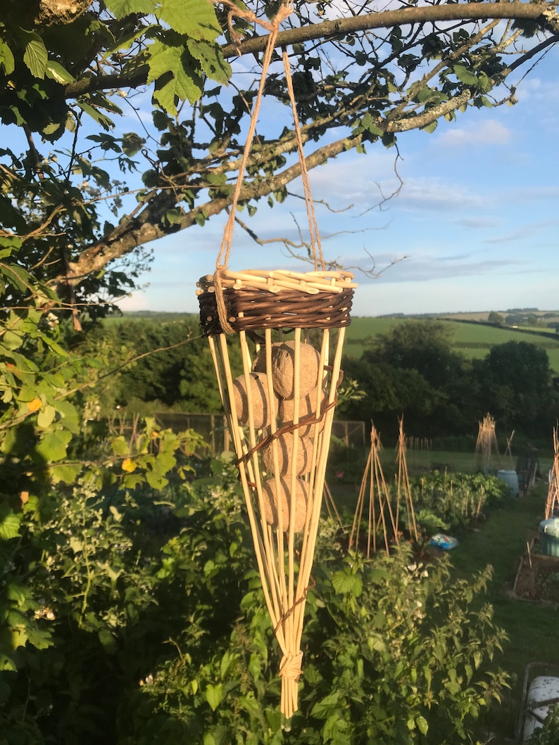 May include: A woven wicker basket with a brown and natural finish hangs from a tree branch. The basket is filled with round, beige balls. The basket is hanging in a garden setting.